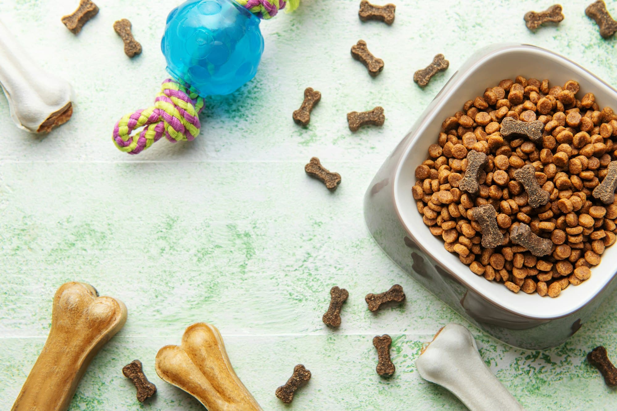 A bowl with dog food, dog treats and toys on a wooden floor.