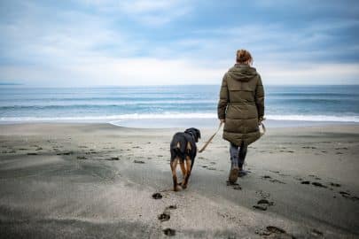 A girl walks with a Rottweiler dog on a leash along the beach in cold weather